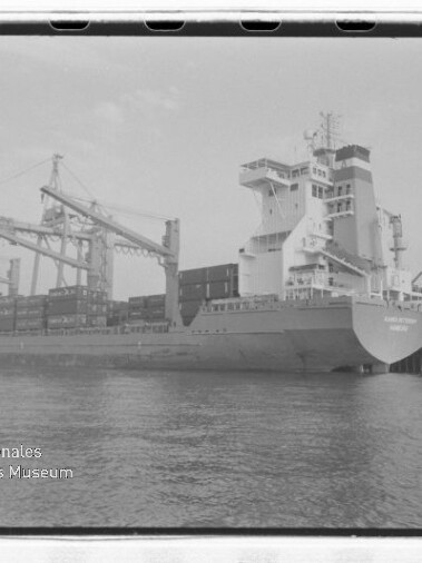 Containerschiff am Kai mit Containern an Deck und großen Hafenkränen im Hintergrund; Museumslogo links unten.