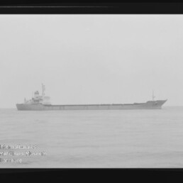 Langes Frachtschiff in Seitenansicht auf ruhiger See, niedrige Ladefläche, Brücke hinten, sichtbarer schwarzer Filmrand.