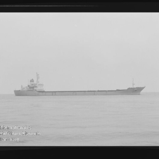 Langes Frachtschiff in Seitenansicht auf ruhiger See, niedrige Ladefläche, Brücke hinten, sichtbarer schwarzer Filmrand.