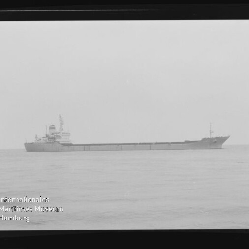 Schwarzweißaufnahme eines langen Frachtschiffs in Seitenansicht auf ruhiger See, mit sichtbarem Filmrähmen und dem Wasserzeichen des Internationalen Maritimen Museums Hamburg in der linken unteren Ecke.