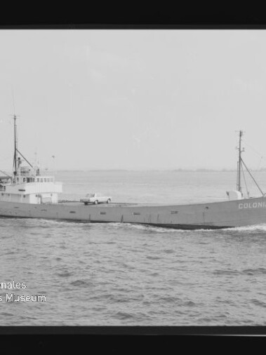 Schwarzweißaufnahme eines Frachtschiffs namens COLONIA, das auf ruhiger See fährt; flacher Rumpf mit aufgesetzten Aufbauten, zwei Masten und leichter Bugwelle, sichtbarer Horizont und Museumswasserzeichen unten links.