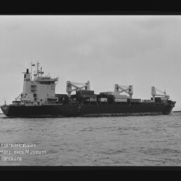 Schwarzweiß-Foto eines Containerschiffs in Seitenansicht auf ruhiger See mit gestapelten Containern, drei Ladekränen auf dem Mitteldeck, Brückenaufbau vorn, sichtbarem Filmrand und dem Wasserzeichen 'Internationales Maritimes Museum Hamburg' unten links.