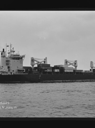 Schwarzweiß-Foto eines Containerschiffs in Seitenansicht auf ruhiger See mit gestapelten Containern, drei Ladekränen auf dem Mitteldeck, Brückenaufbau vorn, sichtbarem Filmrand und dem Wasserzeichen 'Internationales Maritimes Museum Hamburg' unten links.