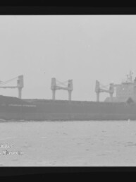 Graustufiges Foto eines Massengutfrachters mit drei großen Deckskränen und achterlicher Brücke, fahrend auf ruhiger See; unten links sichtbares Wasserzeichen des Internationalen Maritimen Museums Hamburg.