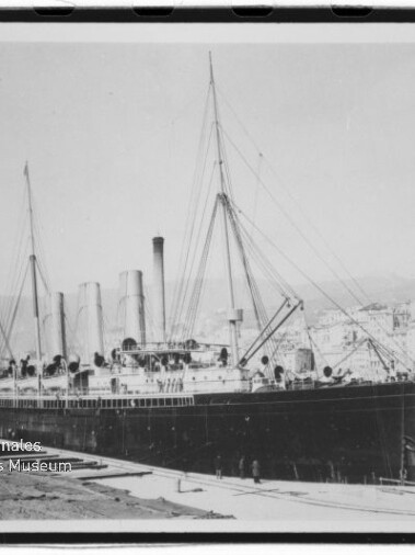 Schwarzweißaufnahme eines großen frühen Ozeandampfers mit mehreren Schornsteinen und Masten, am Kai vor einer Stadt an einem Hügel festgemacht; im Vordergrund Kai und Personen, links unten Wasserzeichen des Internationalen Maritimen Museums Hamburg.