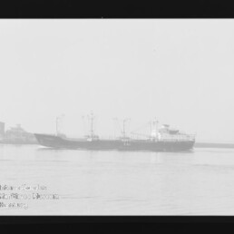 Schwarz-weiß-Foto eines Frachtschiffs mit mehreren Ladekränen, das auf ruhigem Wasser an einem Hafen mit Industriegebäuden vorbeifährt.