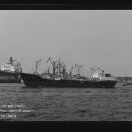 Schwarzweißaufnahme eines Frachtschiffs mit zahlreichen Ladekränen, das im Hafen vor einer Kaianlage liegt, mit leicht bewegter See im Vordergrund und Wasserzeichen des Internationalen Maritimen Museums Hamburg.