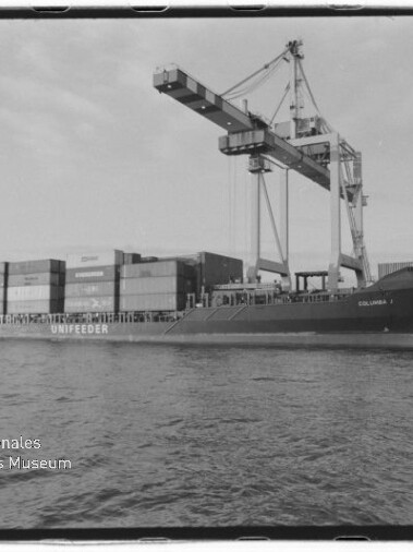 Schwarzweiß-Foto eines Containerfrachters (mit dem Schriftzug UNIFEEDER) am Kai eines Industriehafens, beladen mit gestapelten Containern und von einem großen Portalkran über ihm bedient, mit ruhiger Wasserfläche im Vordergrund und Museumslogo unten links.