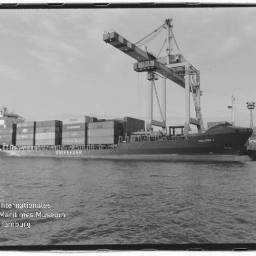 Schwarzweiß-Foto eines Containerfrachters (mit dem Schriftzug UNIFEEDER) am Kai eines Industriehafens, beladen mit gestapelten Containern und von einem großen Portalkran über ihm bedient, mit ruhiger Wasserfläche im Vordergrund und Museumslogo unten links.