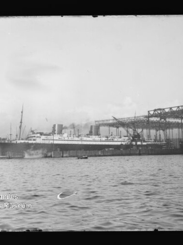 Schwarzweißaufnahme eines großen Passagierschiffs mit mehreren Schornsteinen, im Hafen neben Werftkränen und Stahlgerüsten vertäut, mit Wasser im Vordergrund und Wasserzeichen des Internationalen Maritimen Museums Hamburg unten links.