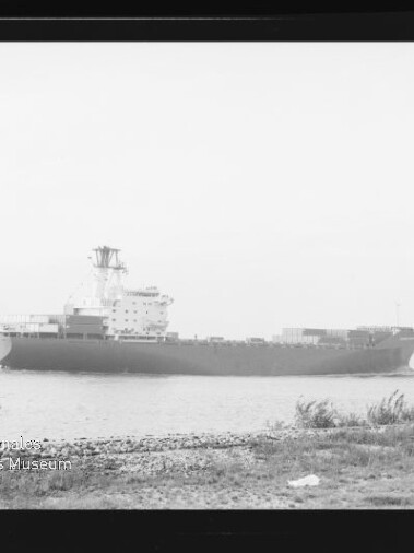 Schwarzweißfoto eines großen Frachtschiffs auf ruhigem Wasser nahe dem Ufer, mit Kiesstrand und Gras im Vordergrund, einem weiteren Schiff in der Ferne und einem Museumslogo unten links.