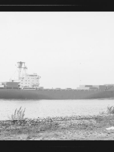 Schwarzweißaufnahme eines großen Containerschiffs in Profilsicht auf ruhiger See, davor ein grasbewachsenes Ufer; unten links sichtbar ein Wasserzeichen/Logo des Internationalen Maritimen Museums Hamburg.