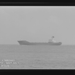 Schwarzweißaufnahme eines weit entfernten Frachtschiffs als dunkle Silhouette auf ruhiger See am Horizont, das Bild ist wie ein Filmnegativ gerahmt; unten links Logo und Schriftzug des Internationalen Maritimen Museums Hamburg.
