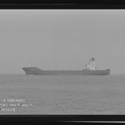 Schwarzweiß-Foto eines seitenansichtigen Frachtschiffs auf ruhiger See, mittig am Horizont, umgeben von sichtbarem Filmrahmen und mit dem Wasserzeichen ‚Internationales Maritimes Museum Hamburg‘ in der linken unteren Ecke.