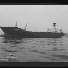 Schwarzweiß-Foto eines großen Frachtschiffs mit der Aufschrift COLUMBUS AUSTRALIS im Hafen, seitliche Ansicht mit mehreren Ladekränen an Deck, ruhige Wasserfläche im Vordergrund und Museumslogo unten links.
