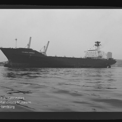 Schwarzweiß-Foto eines großen Frachtschiffs mit der Aufschrift COLUMBUS AUSTRALIS im Hafen, seitliche Ansicht mit mehreren Ladekränen an Deck, ruhige Wasserfläche im Vordergrund und Museumslogo unten links.
