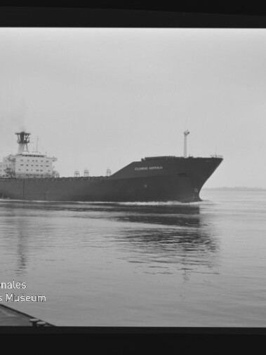 Schwarzweißaufnahme eines großen Frachtschiffs mit dem Namen „Columbus Australia“ auf ruhigem Wasser, leichte Spiegelung im Vordergrund, weiter flacher Horizont und Museumswasserzeichen unten links.