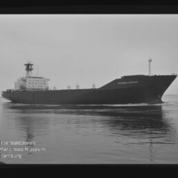 Schwarzweißaufnahme eines großen Frachtschiffs „Columbus Australia“, seitlich vorbeifahrend auf ruhiger See vor flachem Horizont; unten links das Wasserzeichen des Internationalen Maritimen Museums Hamburg.
