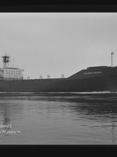 Schwarzweißaufnahme eines großen Frachtschiffs „Columbus Australia“, seitlich vorbeifahrend auf ruhiger See vor flachem Horizont; unten links das Wasserzeichen des Internationalen Maritimen Museums Hamburg.