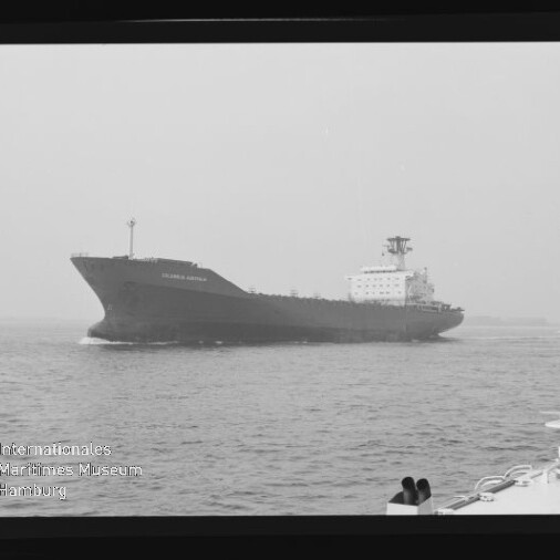 Schwarzweißaufnahme eines großen Frachtschiffs, das in ruhiger See von links nach rechts fährt; rechts im Vordergrund ein Teil eines kleinen Bootes, links unten das Logo des Internationalen Maritimen Museums und am Bildrand der Filmstreifen mit der Aufschrift AGFA PAN 100.