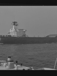 Seitliche Ansicht eines großen Frachtschiffs auf ruhiger See, im Vordergrund Reling und Navigationsgeräte eines anderen Bootes; Schwarzweißaufnahme mit Museumslogo unten links.