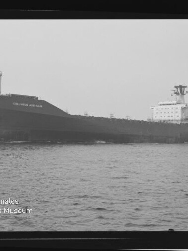 Schwarzweiß-Seitenansicht eines großen Frachtschiffs namens „Columbus Australia“, das ruhig über die See fährt; das Bild ist wie ein Filmnegativ mit Randmarkierungen und Wasserzeichen des Internationalen Maritimen Museums Hamburg gerahmt.