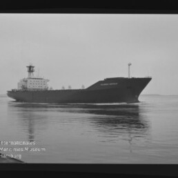 Schwarz-weiß-Foto eines großen Frachtschiffs mit markanter Aufbauten achtern, das bei ruhiger See und bedecktem Himmel über das Wasser fährt, mit Spiegelung im Wasser und dem Logo des Internationalen Maritimen Museums Hamburg unten links.