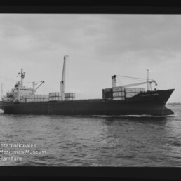 Schwarzweiß-Aufnahme eines Containerschiffs, das mit mehreren auf dem Deck gestapelten Containern und zwei Ladekränen über ruhige See fährt vor flachem Himmel; links unten sichtbar das Wasserzeichen des Internationalen Maritimen Museums Hamburg.