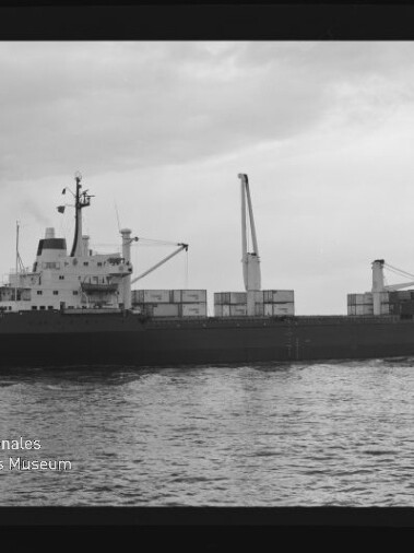 Schwarzweißaufnahme eines Containerschiffs mit gestapelten Containerkästen und einem Ladekran, das bei ruhiger See unter bewölktem Himmel von links nach rechts fährt; unten links sichtbar Logo und Schriftzug des Internationalen Maritimen Museums Hamburg.