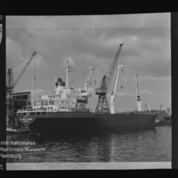 Schwarzweiß-Foto eines im Hafen vertäuten Frachtschiffs neben mehreren großen Hafenkränen, mit Spiegelung im ruhigen Wasser und Wasserzeichen ‚Internationales Maritimes Museum Hamburg‘ links unten.
