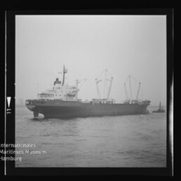 Schwarzweiß-Foto eines Frachtschiffs auf See mit drei sichtbaren Ladekränen und einem kleinen Begleitboot rechts, gerahmt als Filmnegativ mit Logo des Internationalen Maritimen Museums Hamburg.