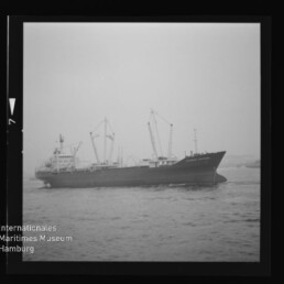 Schwarz-weiß-Foto eines Frachtschiffs mit mehreren Deckskränen vor nebligem Hafen, gerahmt wie ein Fotonegativ und mit Wasserzeichen des Internationalen Maritimen Museums Hamburg unten links.