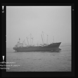 Schwarzweiß-Foto eines Frachtschiffs im Halbprofil mit mehreren Ladekränen auf ruhigem Wasser vor trübem Himmel; das Bild zeigt den Filmnegativ-Rahmen und unten links das Logo ‚Internationales Maritimes Museum Hamburg‘.