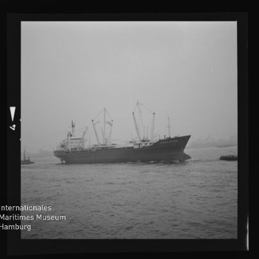 Schwarzweißaufnahme eines Frachtschiffs mit mehreren Deckkränen, das bei trübem, diesigem Wetter in einen Hafen einläuft; links unten das Logo des Internationalen Maritimen Museums Hamburg und der schwarze Filmrand sichtbar.