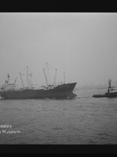 Schwarzweiß-Aufnahme eines Frachtschiffs mit mehreren Ladekränen, das auf dem Wasser von einem Schlepper begleitet wird; im Hintergrund ein nebliger Hafen und am Bildrand Filmkanten sowie das Logo des Internationalen Maritimen Museums Hamburg.