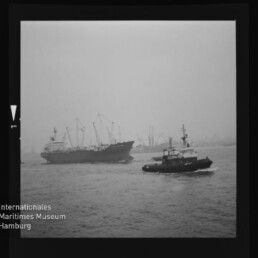 Großes Frachtschiff wird von einem Schlepper begleitet, beide fahren auf ruhigem, nebligem Hafenwasser; im Hintergrund sind Kräne und Hafenanlagen sichtbar, links unten das Wasserzeichen des Internationalen Maritimen Museums Hamburg.