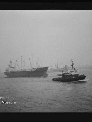 Großes Frachtschiff wird von einem Schlepper begleitet, beide fahren auf ruhigem, nebligem Hafenwasser; im Hintergrund sind Kräne und Hafenanlagen sichtbar, links unten das Wasserzeichen des Internationalen Maritimen Museums Hamburg.