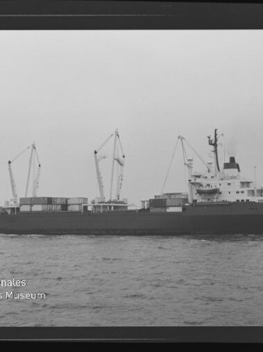 Schwarzweiß-Seitenaufnahme eines Frachtschiffs mit mehreren Ladekränen und Containern, das auf ruhiger See fährt; unten links das Signet ‚Internationales Maritimes Museum Hamburg‘.