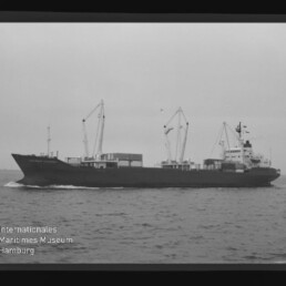 Schwarzweißaufnahme eines Frachtschiffs mit mehreren Deckskränen und auf dem Deck gestapelten Containern, das auf ruhiger See fährt; links unten sichtbar das Wasserzeichen des Internationalen Maritimen Museums Hamburg.