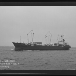 Seitenansicht eines Frachtschiffs mit mehreren Ladekränen, das auf ruhiger See vor flachem Horizont fährt; unten links Wasserzeichen und Schriftzug des Internationalen Maritimen Museums Hamburg.
