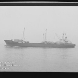 Schwarz-weiß-Fotografie eines Frachtschiffs in Seitenansicht auf ruhiger See mit mehreren Ladekränen an Bord; Filmrand mit 'KODAK'-Schriftzug und Wasserzeichen des Internationalen Maritimen Museums Hamburg.