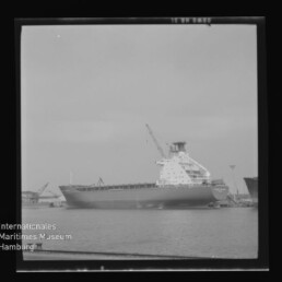 Schwarzweiß-Foto eines großen Frachtschiffs an einem Hafenpier mit aufragendem Aufbau und Kran über ruhiger Wasserfläche, eingefasst in einen quadratischen Negativrahmen; links unten Logo des Internationalen Maritimen Museums Hamburg.