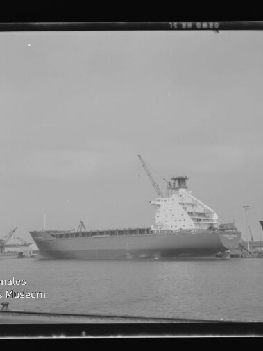 Schwarzweiß-Foto eines großen Frachtschiffs an einem Hafenpier mit aufragendem Aufbau und Kran über ruhiger Wasserfläche, eingefasst in einen quadratischen Negativrahmen; links unten Logo des Internationalen Maritimen Museums Hamburg.