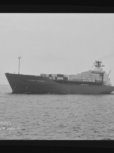 Schwarzweiß-Foto eines Containerschiffs in Seitenansicht mit übereinander gestapelten Containerkästen auf dem Deck, fahrend auf ruhiger See, eingefasst von einem Filmrand und mit dem Logo des Internationalen Maritimen Museums Hamburg unten links.