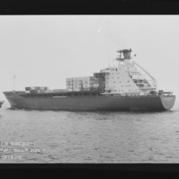 Schwarzweiß-Seitenaufnahme eines Containerschiffs mit gestapelten Containern an Deck, markanten Aufbauten am Heck, ruhiger See im Vordergrund und dem Schriftzug „Internationales Maritimes Museum Hamburg“ unten links.