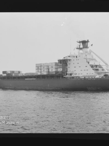 Schwarzweiß-Seitenaufnahme eines Containerschiffs mit gestapelten Containern an Deck, markanten Aufbauten am Heck, ruhiger See im Vordergrund und dem Schriftzug „Internationales Maritimes Museum Hamburg“ unten links.