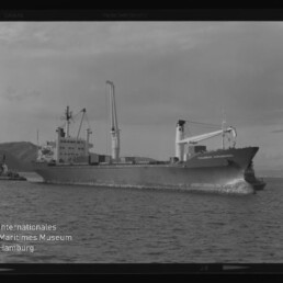 Schwarzweiß-Foto eines großen Frachtschiffs mit Ladekränen und Bugwelle auf ruhiger See, im Hintergrund Küstenhügel und links ein kleines Begleitschiff; Bild zeigt Filmrand und Wasserzeichen des Internationalen Maritimen Museums Hamburg.