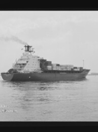 Schwarzweiß-Foto eines Containerschiffs in Seitenansicht auf ruhiger See mit aufgetürmten Containern, rauchendem Schornstein, sichtbarem Filmrand und Museumsstempel in der linken unteren Ecke.