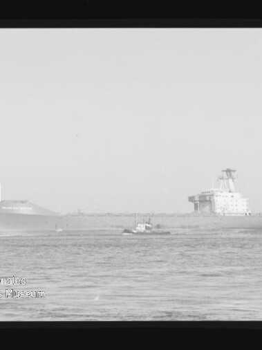 Schwarzweißfoto eines großen Frachtschiffs oder Tankers auf ruhiger See, ein kleines Schlepperboot fährt davor; Bild zeigt Filmrahmen und das Wasserzeichen des Internationalen Maritimen Museums Hamburg unten links.