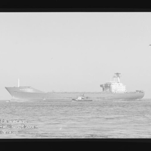 Schwarzweißfoto eines großen Frachtschiffs oder Tankers auf ruhiger See, ein kleines Schlepperboot fährt davor; Bild zeigt Filmrahmen und das Wasserzeichen des Internationalen Maritimen Museums Hamburg unten links.